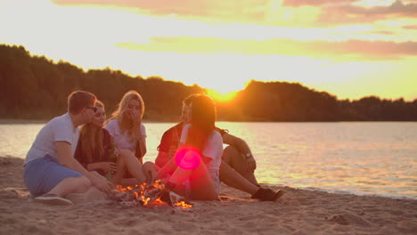 The-young-blonde-is-telling-a-story-to-her-friends-around-bonfire-on-the-beach.-They-are-drinking-beer-at-sunset-and-enjoying-the-summer-evening-on-the-lake-coast.